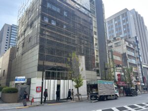 Tall building under construction with metal scaffolding along its facade, street with pedestrians and a delivery truck.