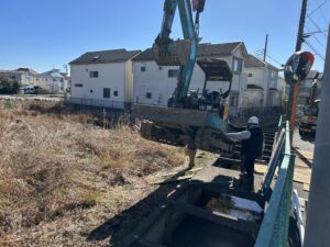 Construction excavator lifts a bucket over a deep trench beside a row of suburban houses, with a worker nearby guiding the operation.