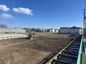 Construction site with a small excavator on dirt, greenhouses to the left, and houses in the background under a blue sky