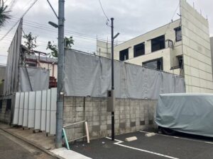 Construction site behind gray tarp fencing with a partially built white building in the background; a large vehicle is covered in the foreground.
