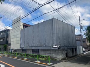 Construction site wrapped in gray tarps and scaffolding along a city street with green railing along the curb.