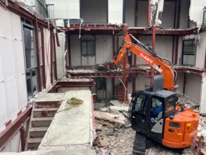 Orange hydraulic excavator operating at a partially demolished three-story building, exposing red steel framing, debris, and exposed interior rooms.