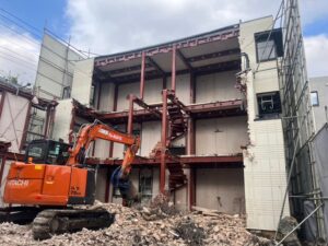 Orange Hitachi excavator at a demolition site, tearing down a multilevel structure with exposed red steel beams and scattered rubble in the foreground.