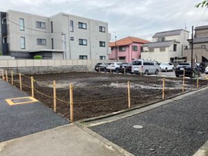 Vacant dirt lot fenced with wooden posts and yellow rope, in a residential street with parked cars and multi-story buildings behind it.