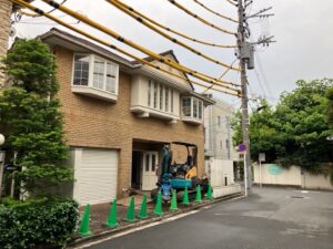 Construction scene: small blue and yellow excavator parked by a brick building, green cones line the sidewalk, utility poles with overhead cables on a quiet street
