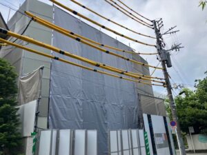 Construction site with a building wrapped in gray protective tarps, metal fencing, and overhead power lines with yellow cables