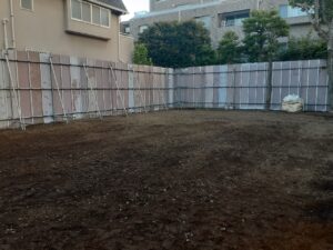 Empty dirt lot bordered by temporary metal fencing and scaffolding panels, with a bag of materials on the right and buildings in the background.