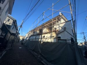 Narrow urban street with a building under construction, surrounded by metal scaffolding and a dark barrier fence, under a clear blue sky.