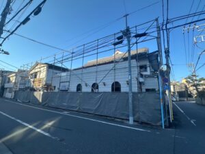 White-tiled building under renovation surrounded by scaffolding and gray fencing on a quiet urban street, with many overhead power lines and a clear blue sky.