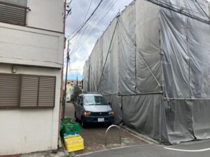 Narrow street with a construction site wrapped in grey tarps beside a white building; a silver van parked at the curb and plastic crates on the sidewalk.