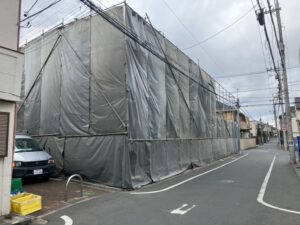 Construction scaffold wrapped in gray tarpaulin covers a building along a narrow residential street, with overhead power lines and a parked van nearby.