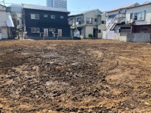 A vacant dirt lot in a residential neighborhood, with brown soil in the foreground and a row of houses with balconies in the background.