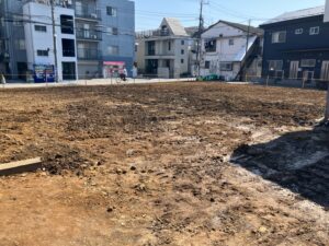 Urban construction site: a bare dirt lot bordered by a sidewalk and low fencing, with apartment buildings across the street and a cyclist riding by on the sidewalk in the background.