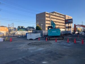 Construction site with a blue Kobelco excavator and an orange excavator, safety cones, and a brick apartment building in the background.