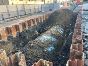 Trench at a construction site with steel sheet piles along both sides and a large dirt mound in the center