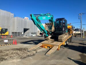 turquoise excavator being loaded onto a flatbed trailer at a construction site with a gray building under wrap and orange cones nearby