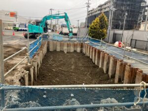 Excavation pit with vertical sheet piles and a turquoise excavator in the background at an urban construction site.