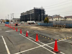 Construction zone in a parking lot with orange cones forming a barrier and caution tape, a white truck nearby and a building under scaffolding in the background.