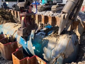Excavator in a trench breaking a cracked blue pipe, with wooden shoring along the edge and workers in the background.