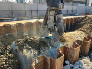 Excavator bucket digging into soil beside a trenches lined with steel sheet piles at a construction site