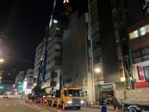 Nighttime construction scene: a crane truck lifts a worker in a bucket beside a tall building, orange cones line the street.