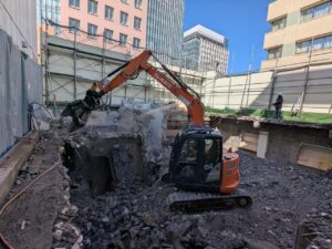 Mini orange excavator with a hydraulic breaker breaking concrete in a rubble-filled construction pit, with scaffolding and a worker on the far edge.