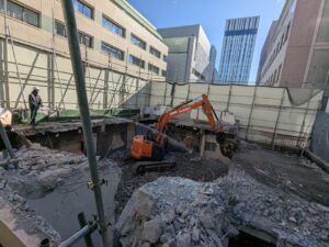 Orange excavator demolishing a concrete building, rubble and rebar scattered, worker in hard hat on left, city skyscrapers in the background.