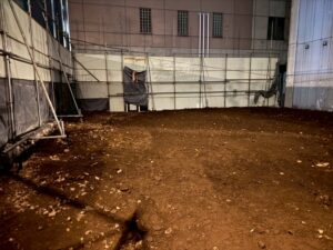 Indoor construction area with bare dirt floor and scattered rocks, surrounded by metal scaffolding and plastic sheeting walls at night.
