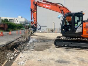 Excavator with a hydraulic breaker breaking up asphalt at a construction site, smoke rising from the rubble and orange safety barriers in the background