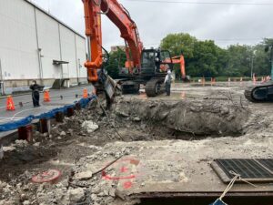 Orange excavator lifting concrete rubble in a deep trench at a construction site, workers in hard hats nearby.