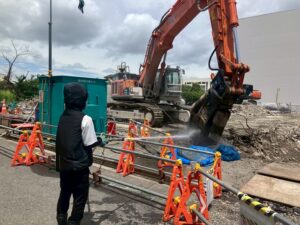 Construction worker in a hooded jacket stands behind orange barricades, using hoses as a large orange excavator pours water onto rubble at a construction site.