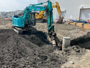 Teal hydraulic excavator at a construction site digging a trench beside a concrete pipe and dirt mound.