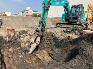 teal excavator with large bucket digging a deep trench at a construction site; worker in helmet watches nearby