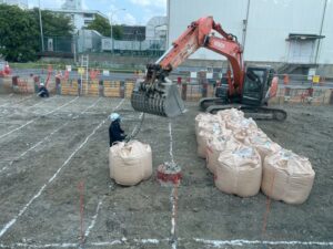Orange Hitachi excavator lifting a large beige bag at a construction site, with workers in hard hats and rows of bags on dirt.