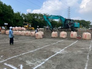 Construction site with a row of large numbered beige sandbags, a teal excavator on a dirt mound, and workers wearing hard hats nearby.