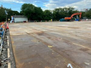 Wide view of a construction yard with large concrete slabs, orange cones, and an orange excavator in the background on the right.