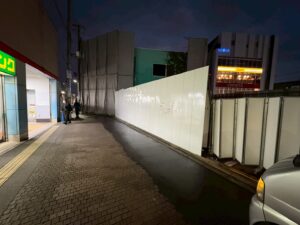 Night street scene at dusk with a tall white construction barrier along the sidewalk, two pedestrians walking, and a storefront with a green sign on the left. A parked van is visible in the foreground on the right.