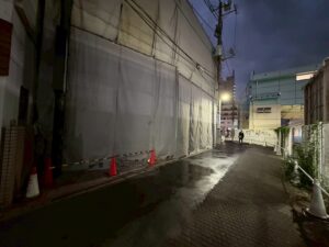 Narrow, wet alley at dusk with a building covered in gray tarps on the left, orange traffic cones and barriers along the curb, and a cyclist in the distance under a streetlight.