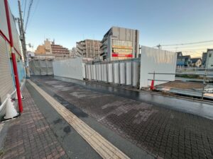 Construction site fenced off along a sidewalk with temporary white panels and metal railing, urban buildings in the background, late afternoon light
