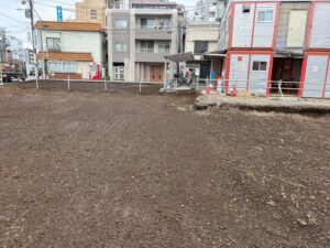Vacant dirt lot with boundary stakes in a city block; row of mixed-use buildings and a red-gray storefront in the background.