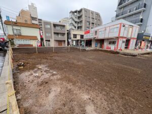Urban construction site: empty, muddy lot fenced off by stakes with surrounding parked buildings and storefronts