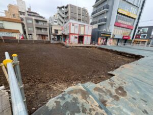 Urban construction site: bare dirt lot bounded by a railing on a city street with multi-story buildings in the background.