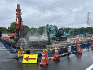 Construction site with orange and teal excavators, orange safety cones, and barrier fencing on a wet, smoky ground under power lines.