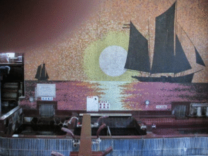 Colorful mosaic mural of sailing ships on a sunset-hued sea, inside a tiled pool area.