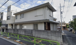 Two-story white urban house with metal siding, fenced front yard, and bright green street railing along a narrow sidewalk under a blue sky with power lines overhead